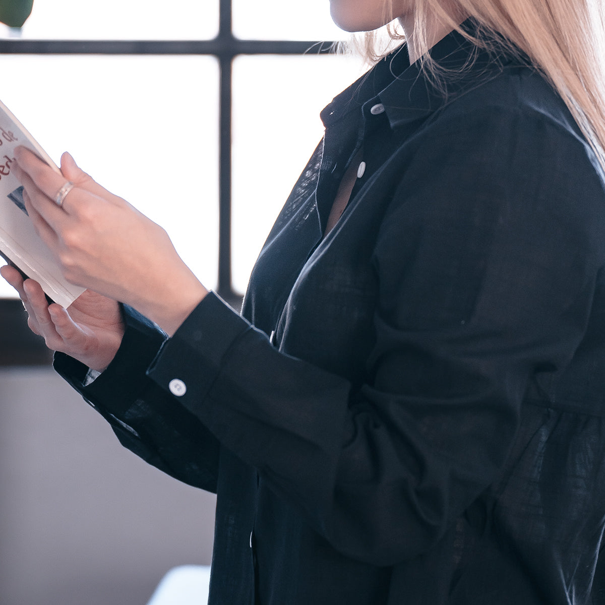 Mujer con camisa negra leyendo cerca de ventana mostrando ropa femenina en línea moderna y elegante